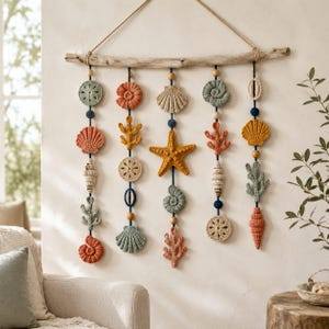 Op de afbeelding: Een wandhanger met vijf verticale strengen opgehangen aan een horizontaal stuk drijfhout. Elke streng toont een reeks gehaakte zeedieren, waaronder schelpen, zeesterren en koraal, afgewisseld met kleine houten en donkerblauwe kralen.