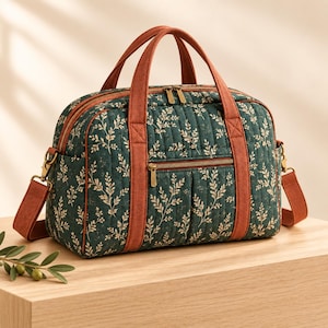 Könnte beinhalten: Eine gesteppte Reisetasche in Dunkelgrün mit einem hellen botanischen Blattmuster. Die Tasche verfügt über rostfarbene Stoffgriffe, passende Besätze und einen abnehmbaren Schultergurt. Sie enthält eine Fronttasche mit Reißverschluss und goldfarbene Beschläge.