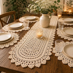 May include: A cream-colored crochet table runner and matching round placemats featuring an intricate lace pattern with scalloped edges. The set is arranged on a wooden table with ceramic plates, clear textured glasses, and a central ceramic vase.