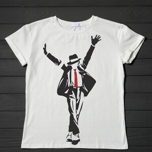 May include: White t-shirt featuring a black silhouette of a person in a hat, suit, and red tie, with arms raised. The design is a stylized illustration, likely a tribute to a famous performer. The shirt is made of a soft material.