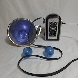 May include: A black and silver Kodak Duaflex II camera with a large circular metal flash reflector, a black cord, and two blue glass flash bulbs. The camera front features the text Kodak Duaflex II Camera and Kodet Lens in white and red lettering.