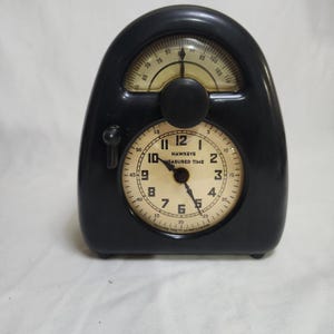 Peut inclure: Un minuteur noir Hawkeye Measured Time avec un cadran crème comportant des chiffres et des aiguilles noirs. L'appareil comprend une jauge arquée supérieure avec une aiguille et un cadran central, ainsi qu'un petit interrupteur latéral ON/OFF.