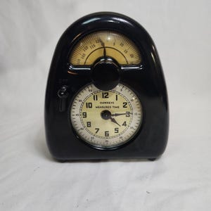 May include: A black Hawkeye Measured Time kitchen timer with a glossy finish. The device features a cream-colored clock face with black numerals and a secondary arched scale at the top ranging from 0 to 120. A black knob and switch are present.