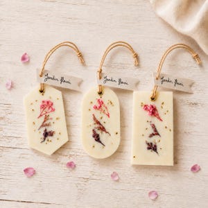Peut inclure: Trois tablettes de cire blanc cassé de formes hexagonale, ovale et rectangulaire, incrustées de fleurs séchées roses et violettes et de flocons dorés. Chaque tablette est suspendue par une ficelle avec une étiquette Jardin fleuri.