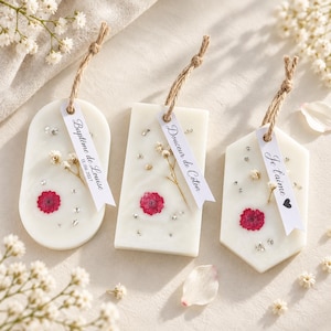 Op de afbeelding: Drie witte wastabletten in ovale, rechthoekige en zeshoekige vormen, elk ingelegd met een klein rood bloemetje, gedroogd wit gipskruid en zilveren glitters. Elk tablet hangt aan een touwtje van jute met een wit papieren label.