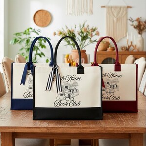 May include: Three rectangular canvas tote bags with solid colored bottoms in navy, black, and burgundy. Each bag features a black line art graphic of a stack of books, a coffee cup, and glasses, with the text "Club Name Book Club" and a ribbon.