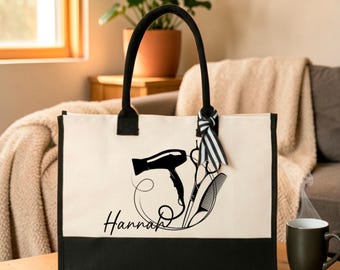 Puede incluir: Un bolso de lona color crema con panel inferior y asas negras. El frente presenta un gráfico negro de un secador de pelo, tijeras y un peine, con el nombre "Hannah" impreso en cursiva negra. Una cinta a rayas blancas y negras está atada a una de las asas.