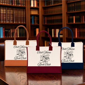 Three tote bags with the text "Book Club" printed on them, placed on a wooden surface in front of a bookshelf.