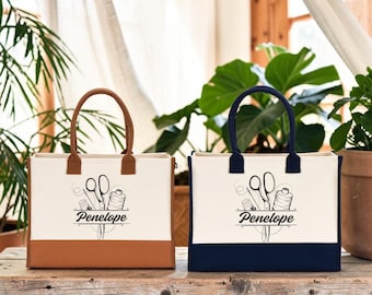 Puede incluir: Dos bolsos tote rectangulares de lona con asas resistentes. Uno tiene base y asas marrones, mientras que el otro tiene base y asas azul marino. Ambos bolsos muestran un gráfico negro de tijeras, un dedal y un hilo con el nombre Penelope.