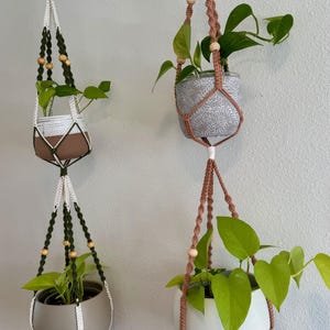 May include: Two macrame plant hangers with potted plants. One hanger is white and green, the other is brown. The pots are gray, brown, and white. The plants have green leaves. The hangers are suspended from a metal bar.