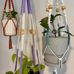 May include: Three macrame plant hangers in shades of brown, purple, and teal, each holding a potted plant. The hangers feature wooden beads and tassels. The pots are white and gray, with green plants inside.