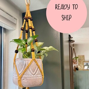 Puede incluir: Un colgador de plantas de macramé amarillo, que sostiene una planta en maceta con hojas verdes. El colgador está adornado con cuentas de color marrón oscuro. La maceta tiene una superficie texturizada de color claro. Las palabras "READY TO SHIP" están en un círculo rosa.