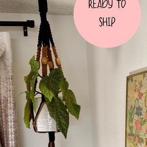 May include: A macrame plant hanger with a white ceramic pot holding a green plant with white spots. The hanger is black and brown, with wooden beads. The text "READY TO SHIP" is in a pink circle.