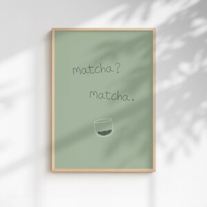 May include: A framed art print with a pale green background and a wooden frame. The print features the handwritten text "matcha? Matcha." below which is a simple illustration of a glass with a dark green substance.