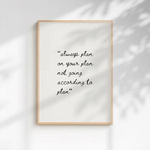 May include: A framed print with the quote "always plan on your plan not going according to plan" in black script on a white background. The frame is a light wood color, and the background has shadows.