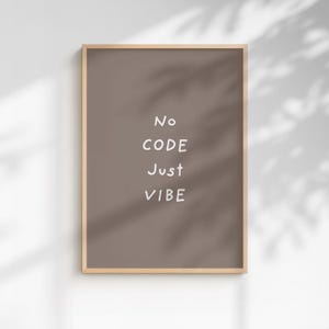 May include: A framed print featuring the words "No CODE Just VIBE" in white on a brown backdrop. The frame is light wood-toned. The print is displayed on a white wall, with shadows cast across it.