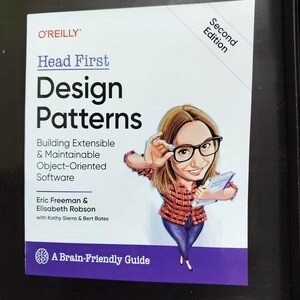 May include: A book titled "Head First Design Patterns" by O'Reilly. The cover shows a cartoon illustration of a person wearing glasses and holding a book. The book's subtitle is "Building Extensible & Maintainable Object-Oriented Software."