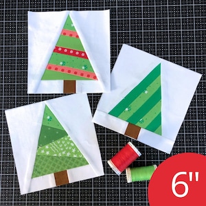 May include: Three square fabric quilt blocks featuring pieced Christmas tree designs on white backgrounds, each measuring 6 inches. The trees are constructed from various green patterned fabrics with brown trunks, accompanied by red and green sewing thread spools.