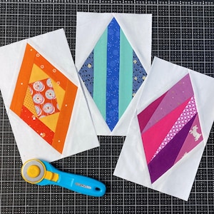 Könnte beinhalten: Drei rechteckige Stoff-Quiltblöcke mit rautenförmigen Mittendesigns in Orange-, Blau- und Pinktönen, begleitet von einem blau-gelben Rollschneider zum Stoffschneiden auf einer schwarzen Schneidematte mit Raster.