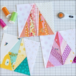 May include: Three quilt blocks in progress, featuring a patchwork design with various patterned fabrics in shades of pink, orange, yellow, and teal. Spools of thread in orange, green, and pink are visible, along with a rotary cutter. The blocks are on a cutting mat.