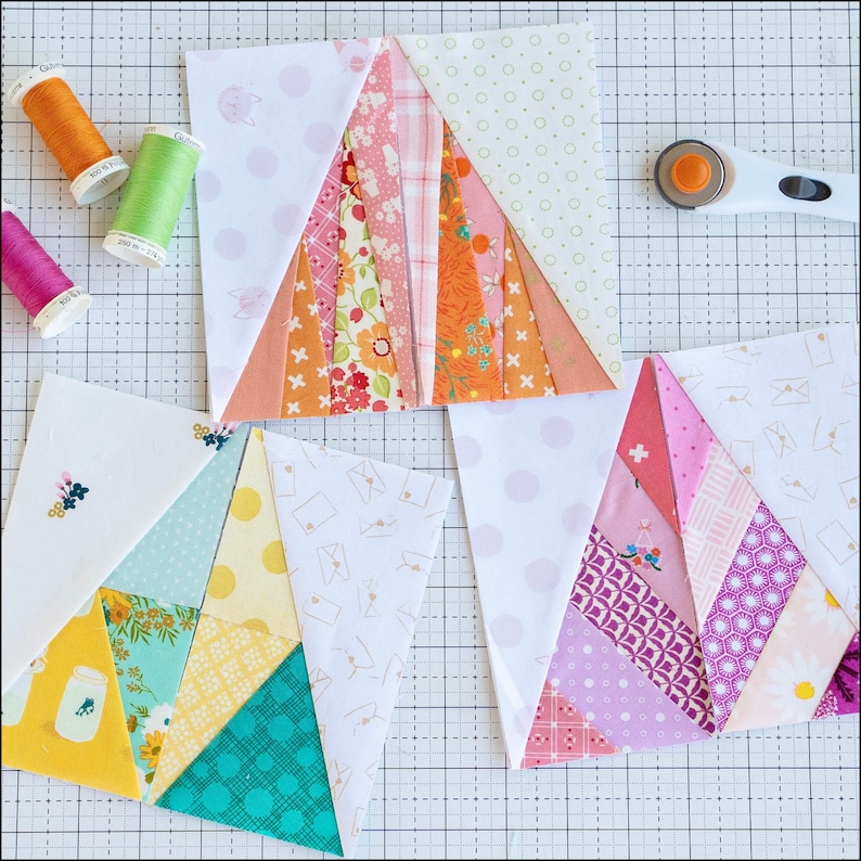May include: Three quilt blocks in progress, featuring a patchwork design with various patterned fabrics in shades of pink, orange, yellow, and teal. Spools of thread in orange, green, and pink are visible, along with a rotary cutter. The blocks are on a cutting mat.