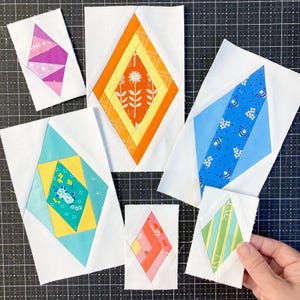 May include: Six rectangular fabric quilt blocks featuring diamond-shaped centers in various colors and patterns, including orange floral, blue bee print, teal floral, and pink geometric designs, all set against a white background fabric.