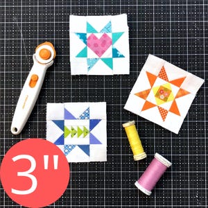 May include: Three colourful quilt blocks with star and heart designs, measuring 7.6 cm. A rotary cutter and spools of yellow and pink thread are also present. The blocks feature various colours including pink, teal, orange, and blue.