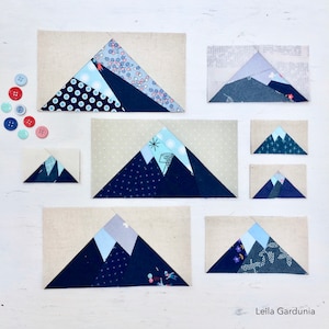 Pode incluir: Um conjunto de nove blocos de montanha de patchwork de tecido em vários tamanhos. Os blocos são feitos de diferentes tecidos com padrões geométricos em tons de azul, cinza e branco. Os blocos estão dispostos sobre uma superfície branca com alguns botões espalhados ao redor.