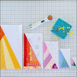 May include: Four different sizes of fabric patchwork quilt blocks, each with a different pattern of colorful fabric triangles. The blocks are arranged in a row on a white gridded cutting mat. A rotary cutter and a pincushion with pins are also visible.