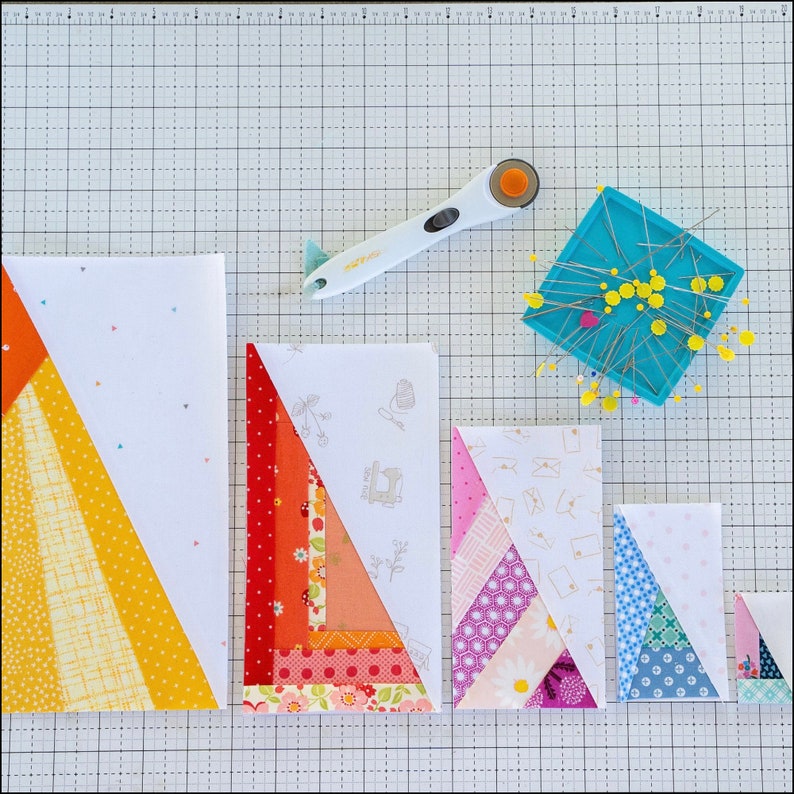 May include: Four different sizes of fabric patchwork quilt blocks, each with a different pattern of colorful fabric triangles. The blocks are arranged in a row on a white gridded cutting mat. A rotary cutter and a pincushion with pins are also visible.