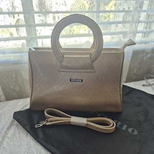 May include: A beige handbag with a textured crocodile-skin pattern, featuring a round top handle and a rectangular metal plaque with the text VITTORIO. The bag includes a detachable, adjustable shoulder strap with silver-toned metal clips.