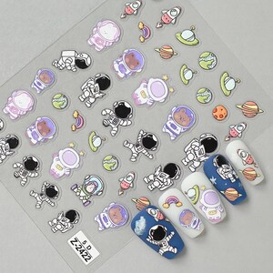 May include: Clear sheet of nail art stickers featuring astronauts, planets, rockets, and rainbows. The stickers include a variety of astronaut designs, some with bears, and are in shades of white, purple, blue, and green. The sheet also includes the text "Z-2422".