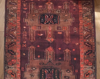 Puede incluir: Una alfombra rectangular con una base de color ciruela profundo y marrón oscuro, con intrincados patrones geométricos en tonos naranja, rojo y crema. El diseño incluye un motivo de medallón central, formas simétricas y un borde detallado.