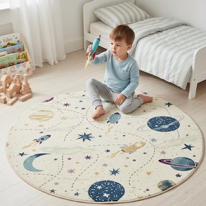Può includere: Tappeto rotondo per bambini a tema spazio. Presenta pianeti, razzi, stelle e una luna crescente in blu, viola e oro. Il tappeto ha un diametro di circa 120 cm.