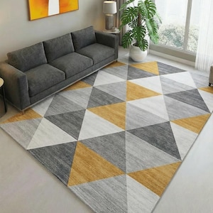 May include: A geometric area rug with a modern design. The rug features a pattern of triangles in shades of gray, white, and gold. The rug is placed in a living room setting with a gray sofa and a large window.