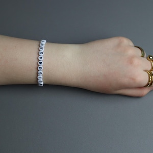 May include: A white beaded bracelet spelling out "INKSVERSIF" is worn on a wrist. The hand is adorned with multiple gold rings. The background is a solid gray.