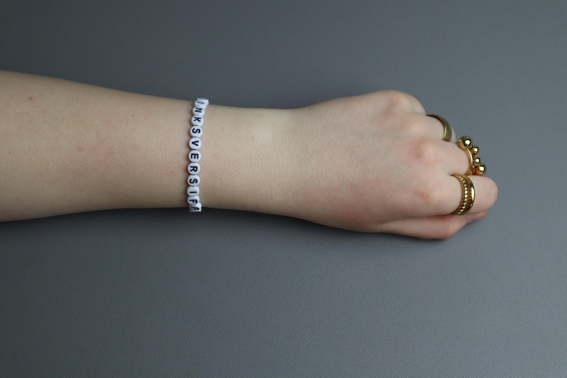 May include: A white beaded bracelet spelling out "INKSVERSIF" is worn on a wrist. The hand is adorned with multiple gold rings. The background is a solid gray.