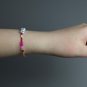 May include: A beaded bracelet with white lettered beads, red and pink beads, and a gold charm. The bracelet is on a wrist, with a gold ring on a finger. The background is a solid gray.