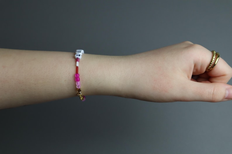 May include: A beaded bracelet with white lettered beads, red and pink beads, and a gold charm. The bracelet is on a wrist, with a gold ring on a finger. The background is a solid gray.