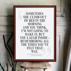 Puede incluir: Un cuadro enmarcado con una cita de Charles Bukowski. El texto, en negrita negra, dice: «A veces te levantas de la cama por la mañana...» El marco es de color marrón oscuro y el fondo es blanco.