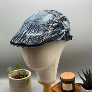 Könnte beinhalten: Eine Jeans-Newsboy-Cap mit verwaschenem, blauem Farbton. Die Cap hat einen gebogenen Schirm und einen verstellbaren Riemen. Die Mütze wird auf einem Schaufensterpuppenkopf präsentiert.