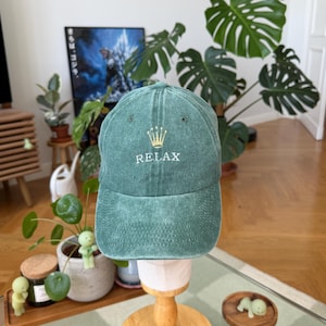 Puede incluir: Una gorra de béisbol de color verde bosque con una textura desgastada y lavada. La parte frontal presenta un icono de corona dorada bordado sobre el texto blanco en mayúsculas que dice RELAX. La gorra tiene una copa estructurada y visera curva.