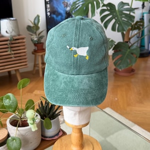 Op de afbeelding: Een saliegroene baseballpet met een verwassen stoffen afwerking. Het voorpaneel is voorzien van een geborduurde afbeelding van een witte gans met gele poten die een klein mes in de snavel houdt. De pet heeft een gestructureerde kroon en klep.