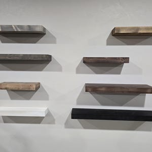 May include: A collection of eight floating shelves in various wood tones, including gray, brown, and black, mounted on a light gray wall. The shelves are rectangular and vary in size, offering a modern storage solution.