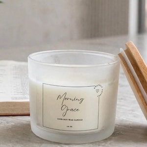 May include: A frosted glass candle jar with a white label featuring the text Morning Grace 100% soy wax candle 16 oz. The candle has a light-colored wax interior and is accompanied by a separate round wooden lid resting on its side next to the jar.