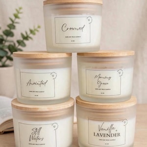 May include: Five frosted glass soy wax candles with light wood lids, stacked in a pyramid. Each candle has a white rectangular label with black text reading Crowned, Anointed, Morning Grace, Still Waters, or Vanilla + Lavender, and 14 oz.