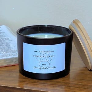 May include: A black cylindrical candle jar with a white label featuring the text Paws of Grace Collection, Paws Play & Pray, Soy Wax, 10 oz, and Heavenly Guided Candles. The candle has a light-colored wax interior and a separate light wood lid.