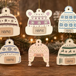 Puede incluir: Seis adornos colgantes de madera con forma de gorros de invierno en varios colores, incluyendo crema, morado, azul, rosa y verde. Cada gorro presenta un patrón único de copos de nieve, corazones o renos e incluye una etiqueta con la palabra Name.