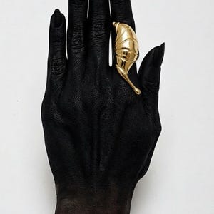 Puede incluir: Un anillo dorado con forma de hoja. El anillo se lleva en una mano pintada de negro con uñas negras. El diseño del anillo presenta vetas detalladas y se coloca sobre un fondo blanco liso. El anillo es una pieza llamativa.
