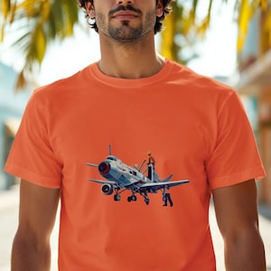 May include: Orange crew neck t-shirt featuring a detailed illustration of a vintage airplane with a pilot and a mechanic. The airplane is primarily blue and silver, with red and black accents. The pilot is wearing an orange helmet.